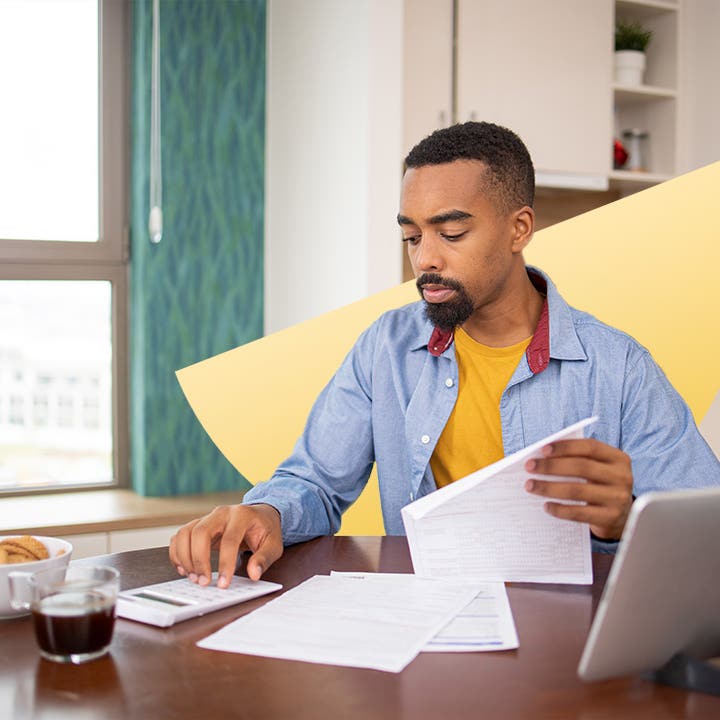 Young Black man uses a calculator at his table while looking through papers.
